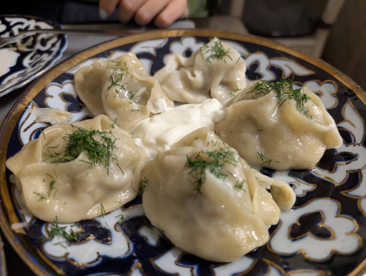 Five large steamed dumplings scattered with chopped dill around a spoonful of soured cream, on a deep blue and gold patterned plate.