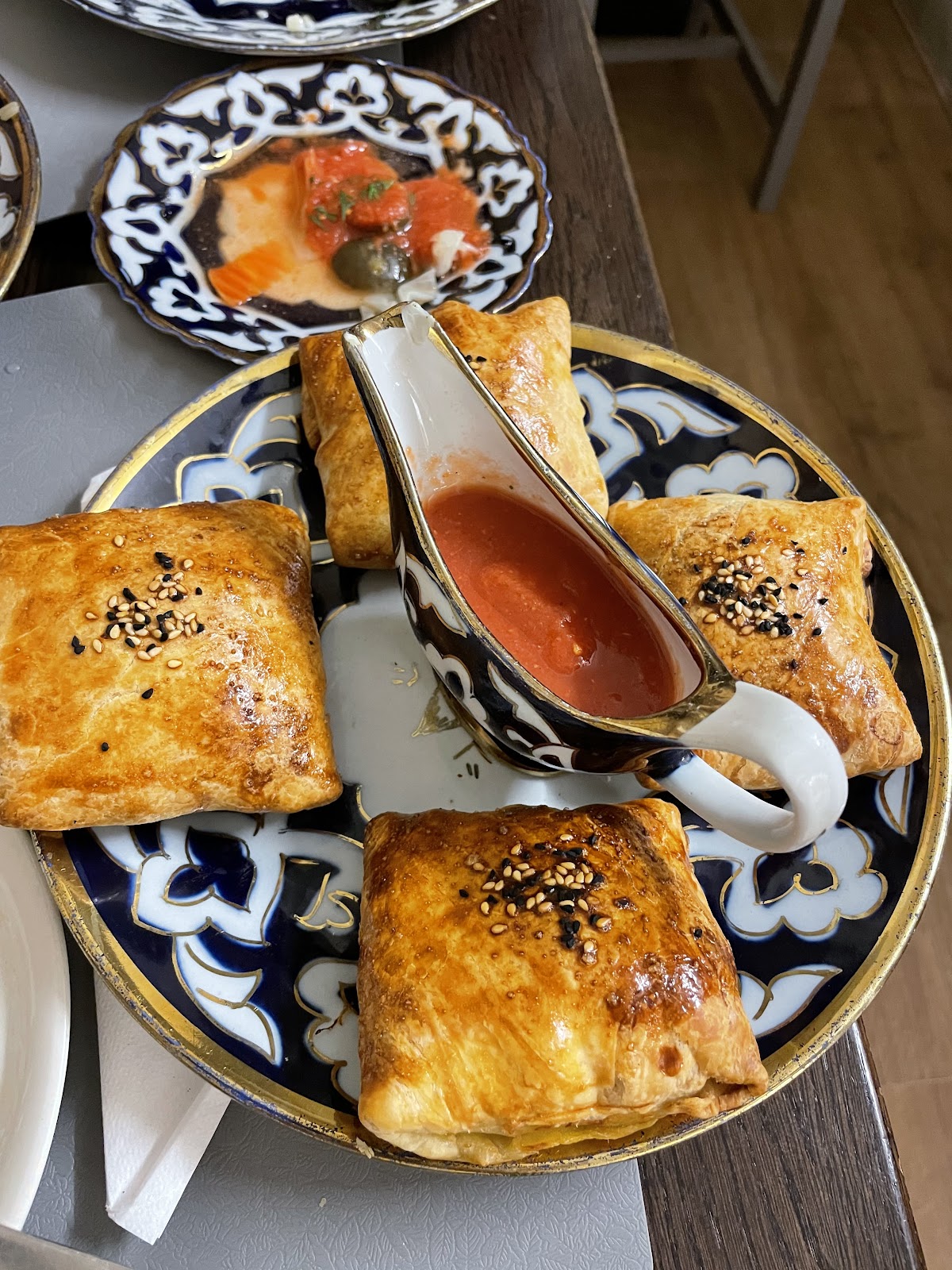 Four square glazed pastries topped with sesame and nigella seeds on a blue and gold plate, with a small patterned jug of red sauce in the middle.