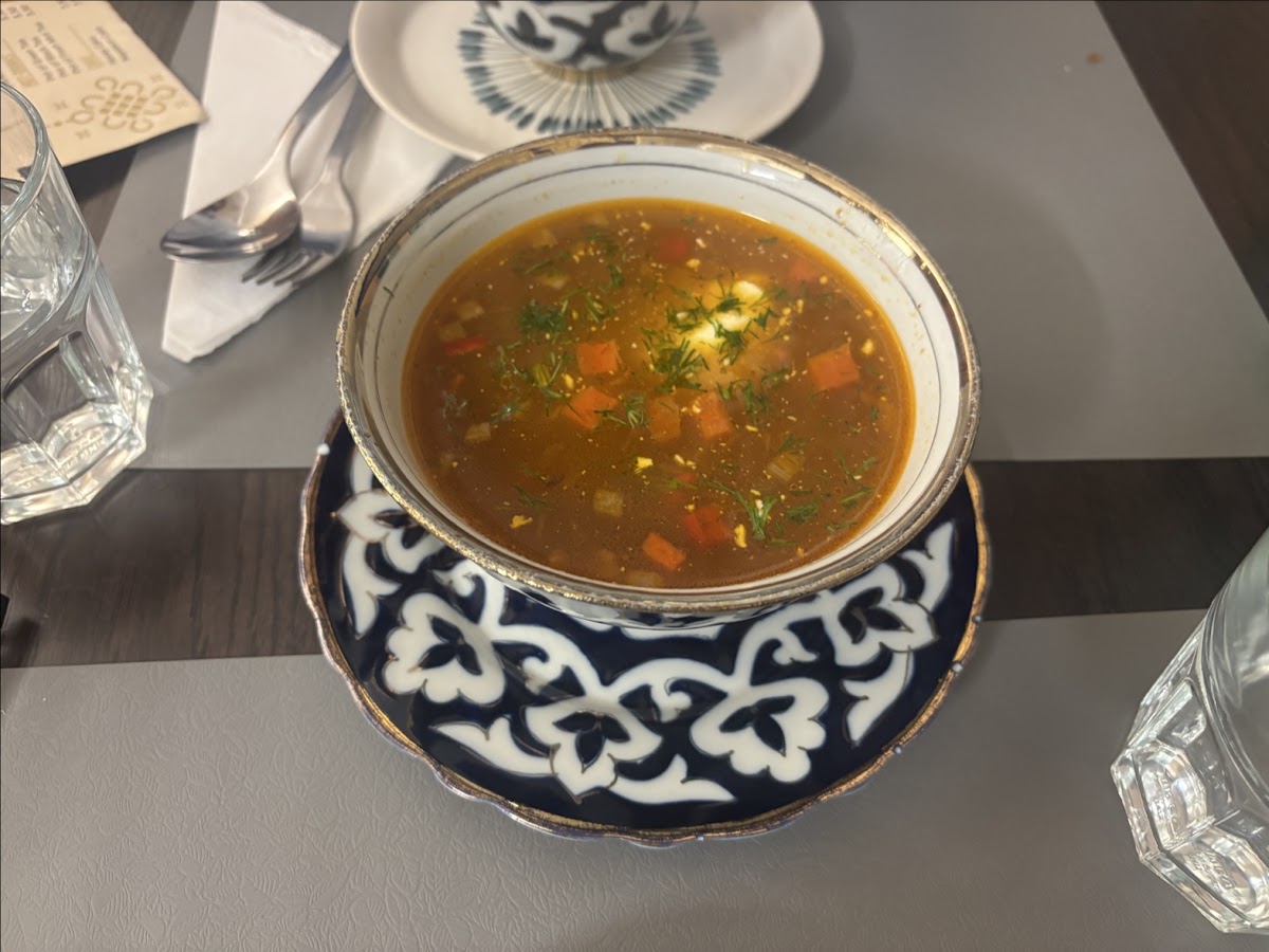 A bowl of clear amber soup with diced vegetables and dill, set on a matching blue, white and gold saucer, with cut-glass tumblers beside it.